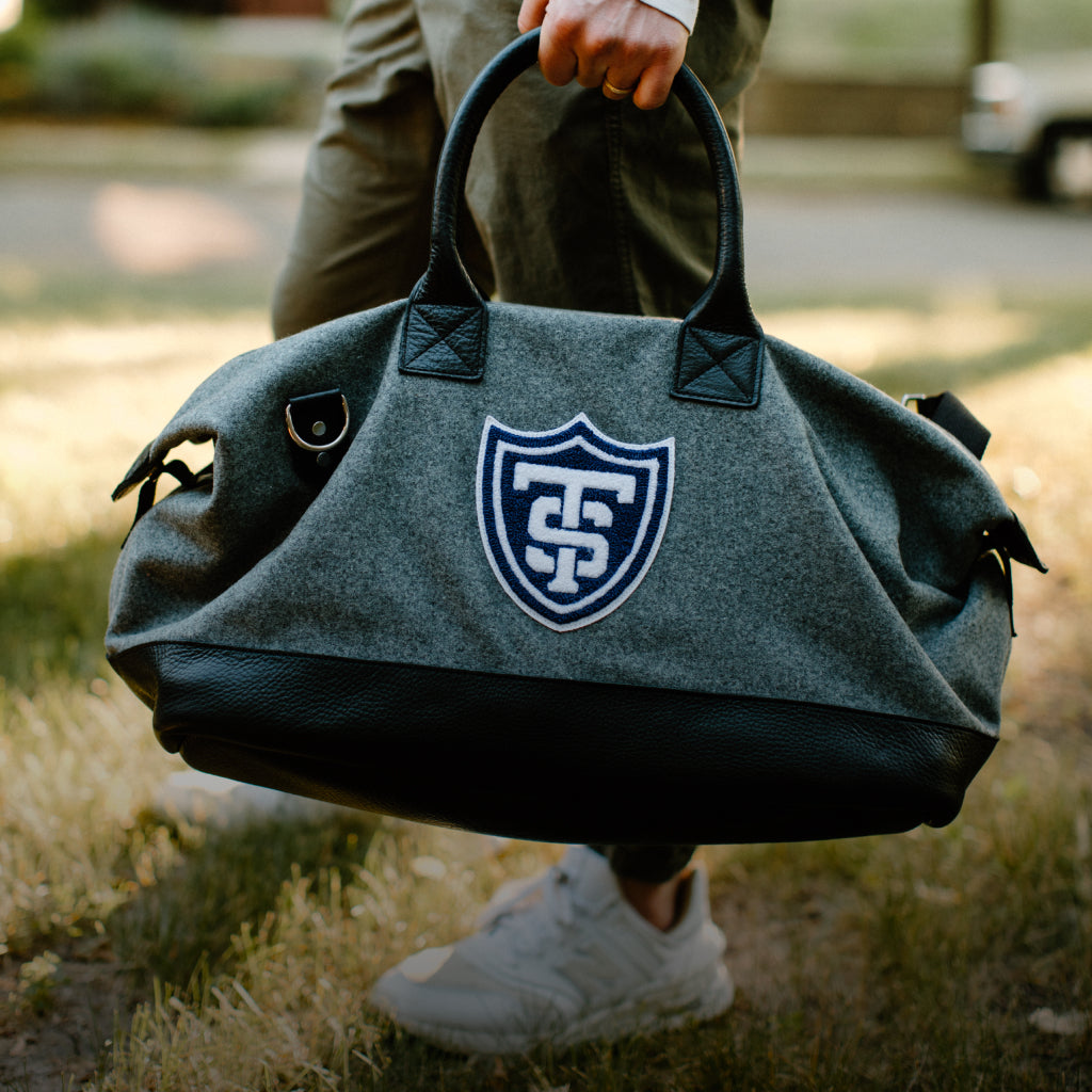close up of man carrying gray University of St. Thomas weekender bag through grass 