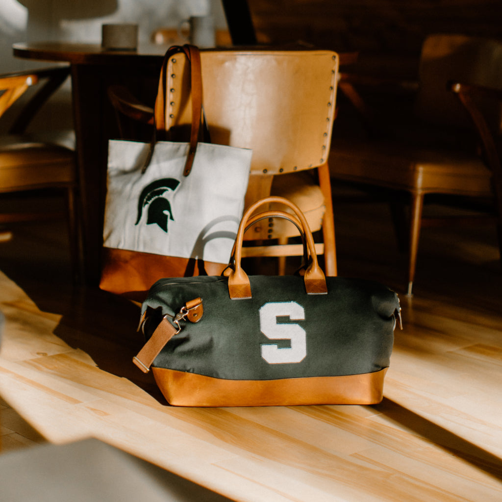 A white tote bag with a Michigan State spartan logo hanging over a chair, below it a green weekender with an S logo